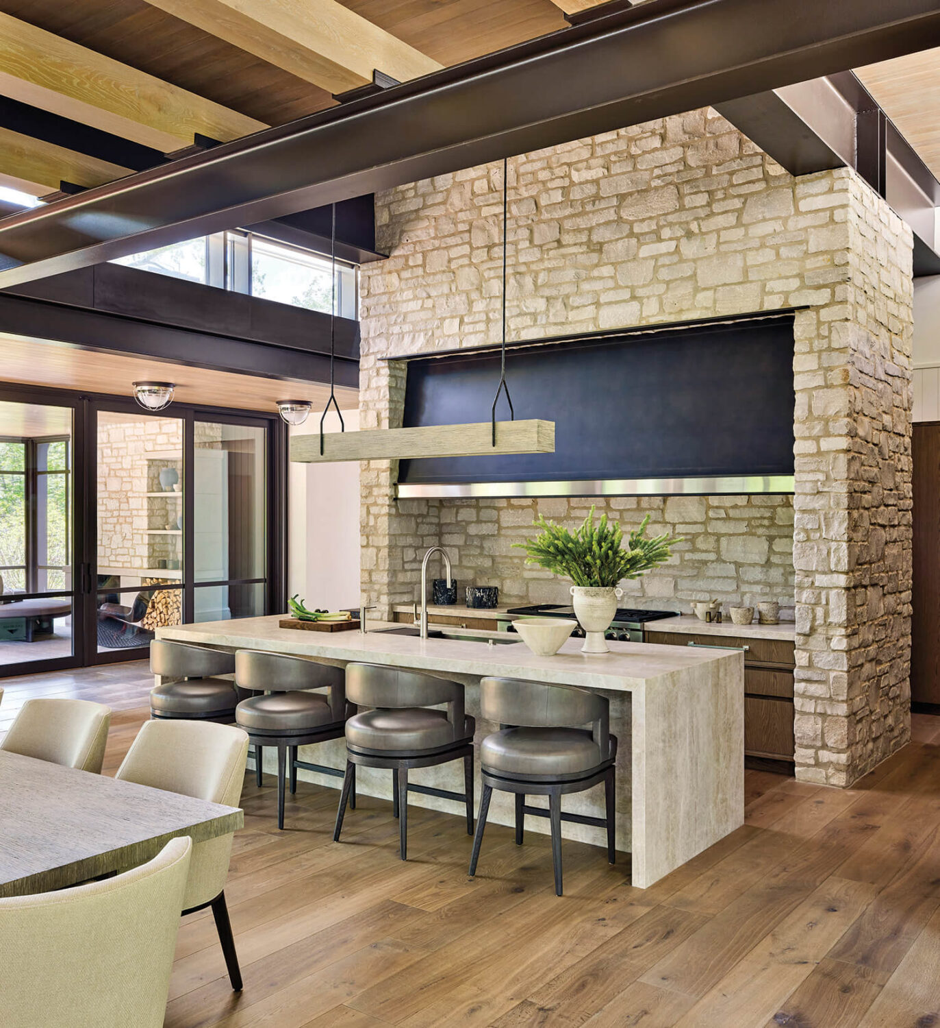 Modern kitchen with stone walls, wood ceiling beams, island with three bar stools, large pendant light, and potted plant, adjacent to dining area and glass doors.