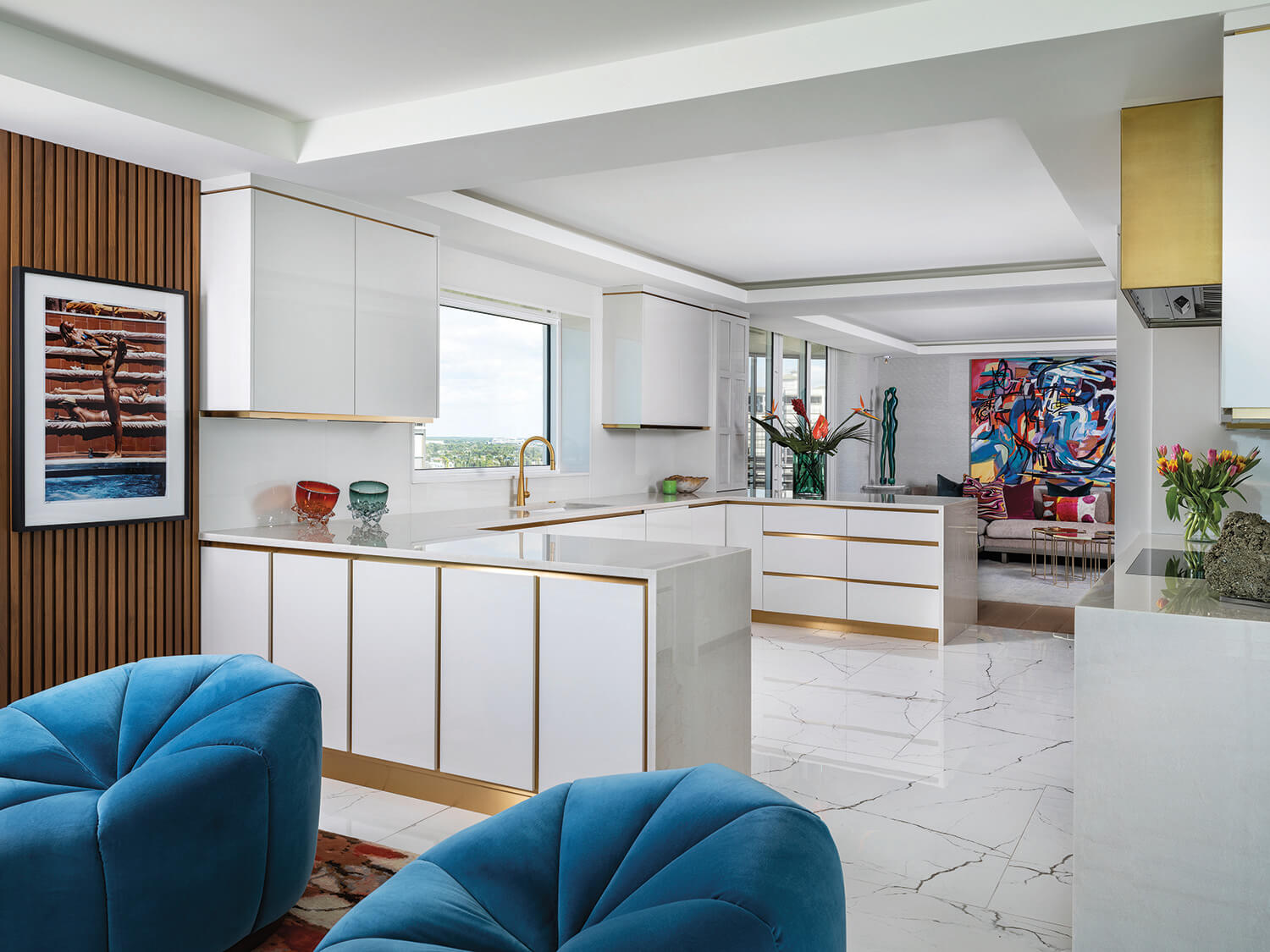 Modern kitchen with white cabinets, gold accents, marble floors, two blue velvet chairs, wall art, and a colorful abstract painting in the adjacent living area.
