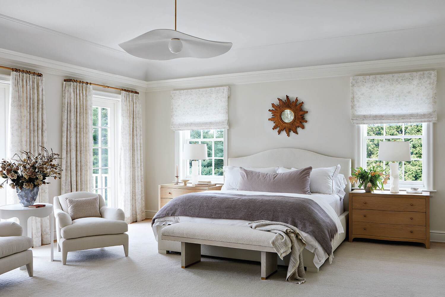 Bright bedroom with a large bed, wooden nightstands, armchairs, light curtains, a sunburst mirror above the bed, and a modern ceiling light fixture.