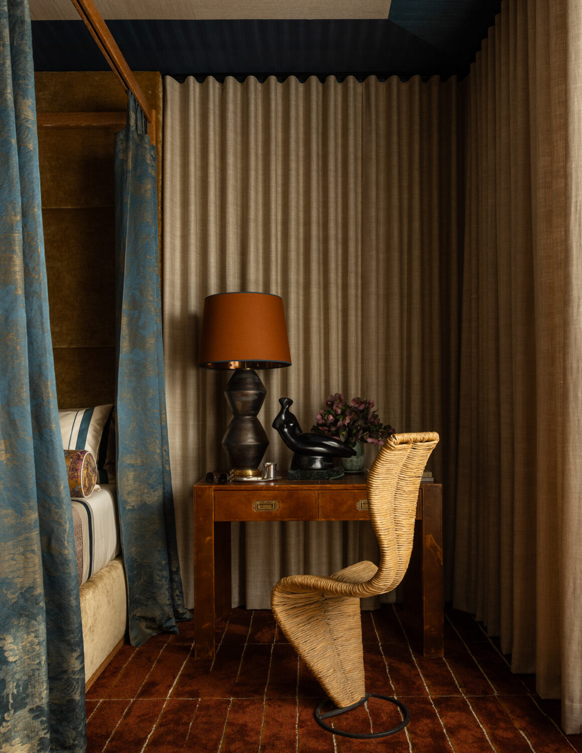 A small brown desk with a lamp, decorative sculpture, and flowers sits by a woven chair in a cozy room with beige curtains and patterned blue bedding.