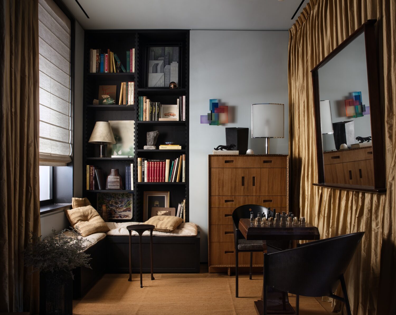 Cozy study with a window seat, built-in black shelves, books, a wooden cabinet, a desk with a chess set, gold curtains, and abstract art reflected in a large mirror.