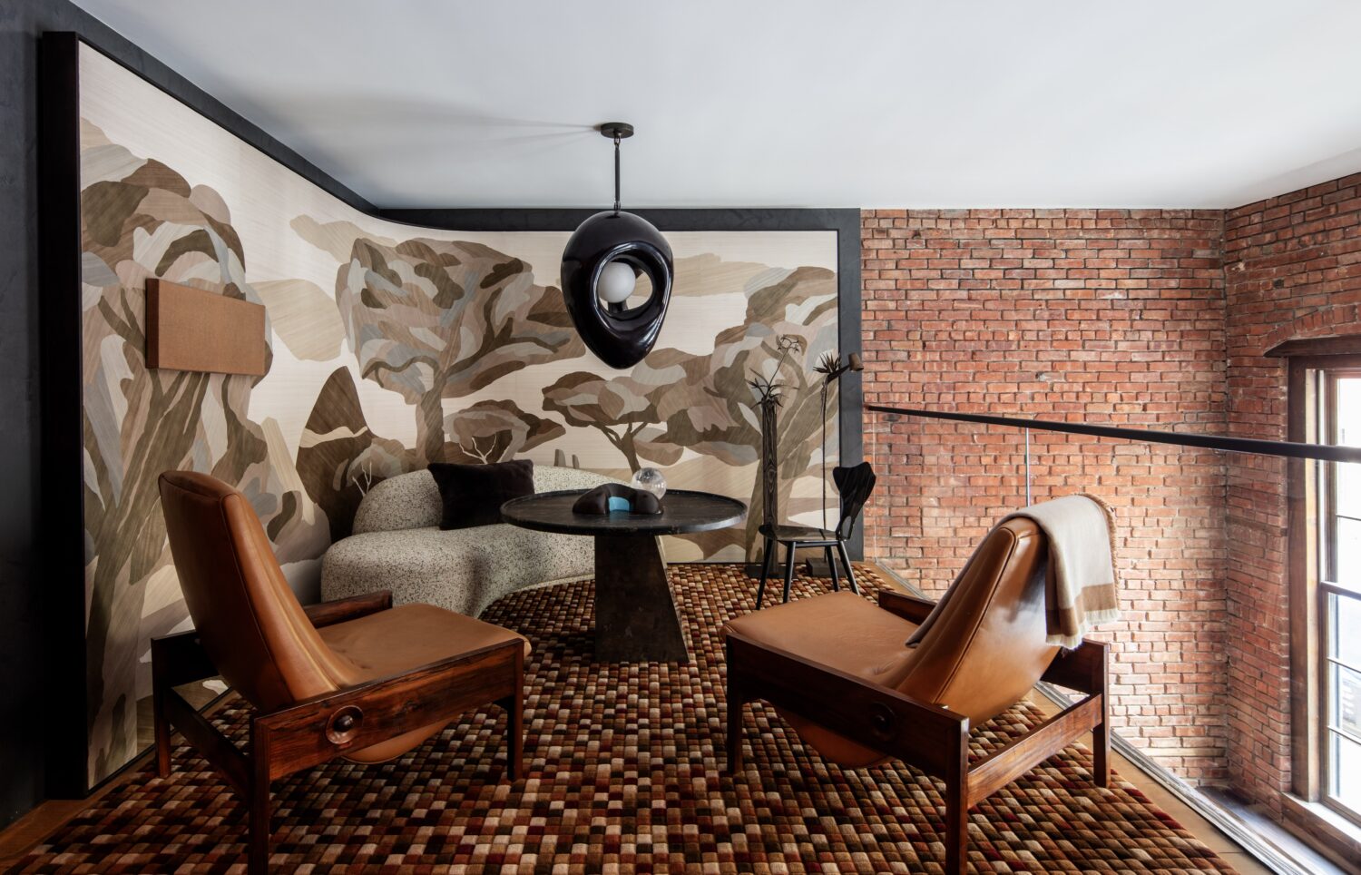 A loft sitting area with brown leather chairs, a round table, abstract wallpaper, a modern black pendant light, and exposed brick walls.