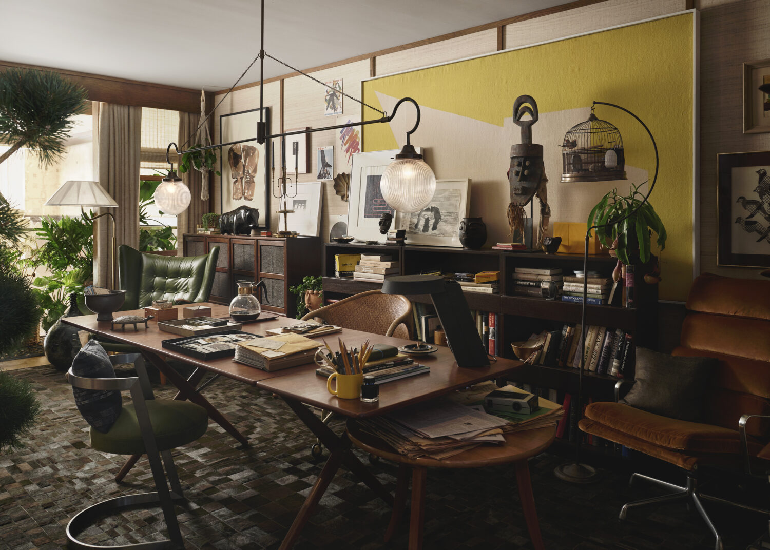 Eclectic home office with wooden desk, various chairs, bookshelves, art, and plants, featuring warm lighting and a mix of decorative objects and work materials.