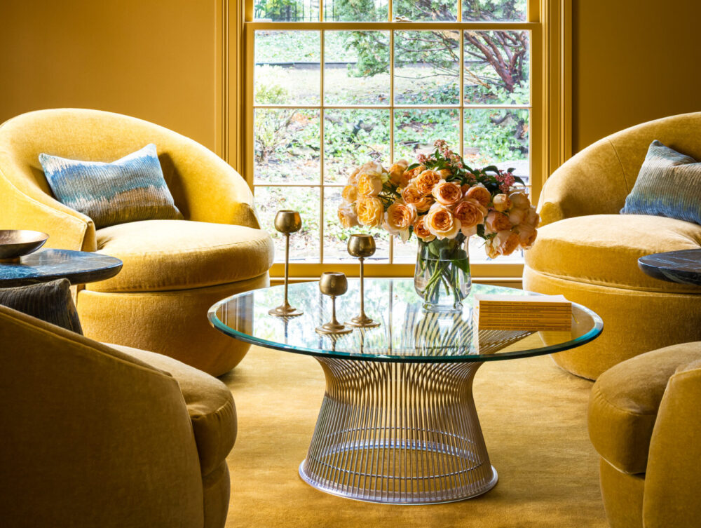 A yellow living room with round chairs, a glass coffee table with flowers, gold decor accents, and a large spherical pendant light, with a window showing greenery outside.