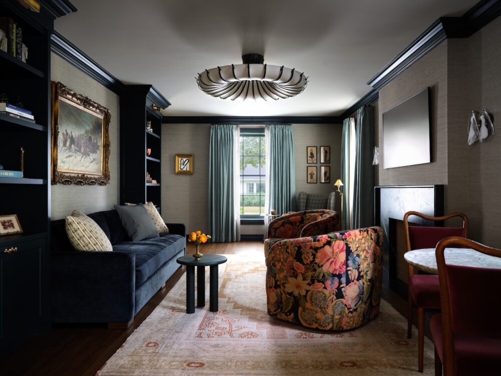 A living room with a dark sofa, floral armchairs, built-in shelves, large window with blue curtains, wall art, and a modern chandelier.