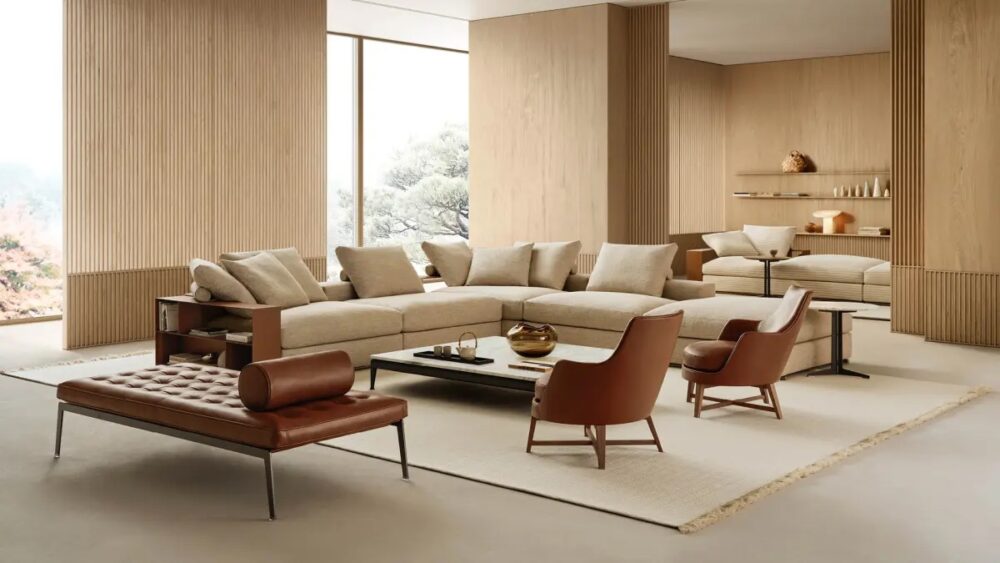 Modern living room with a large beige sectional sofa, two brown leather chairs, a low ottoman, a rectangular coffee table, and wood-paneled walls. Large windows provide natural light.
