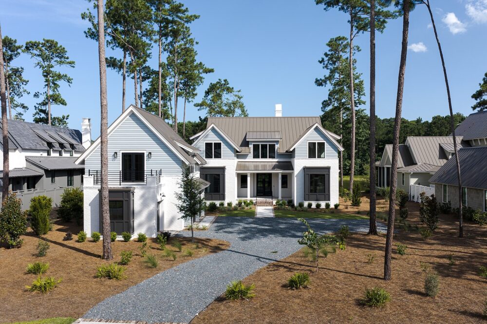 Modern two-story house with gray roof, gravel driveway, and surrounding pine trees under a clear blue sky.