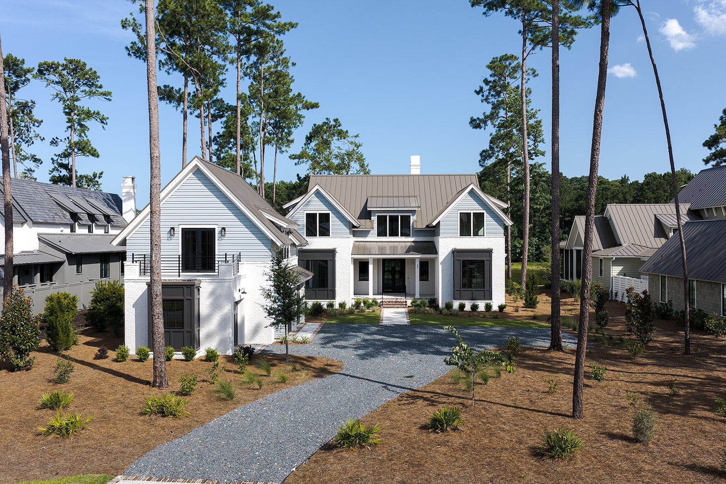 Modern two-story house with gray roof, gravel driveway, and surrounding pine trees under a clear blue sky.