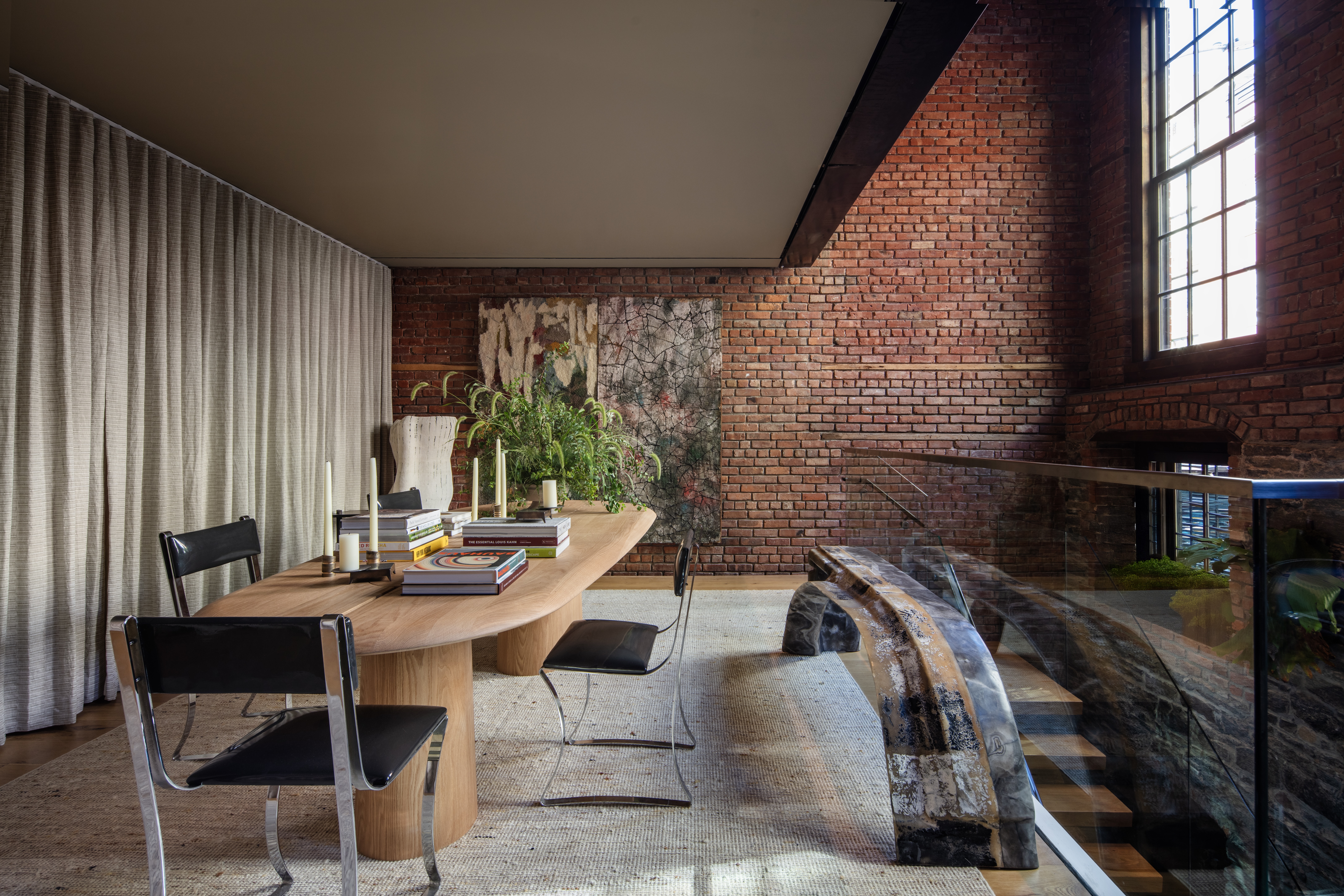 Modern dining room with a wooden table, black chairs, books, plants, a large window, exposed brick walls, and glass railing overlooking an arched brick structure below, by Marmol Radziner.
