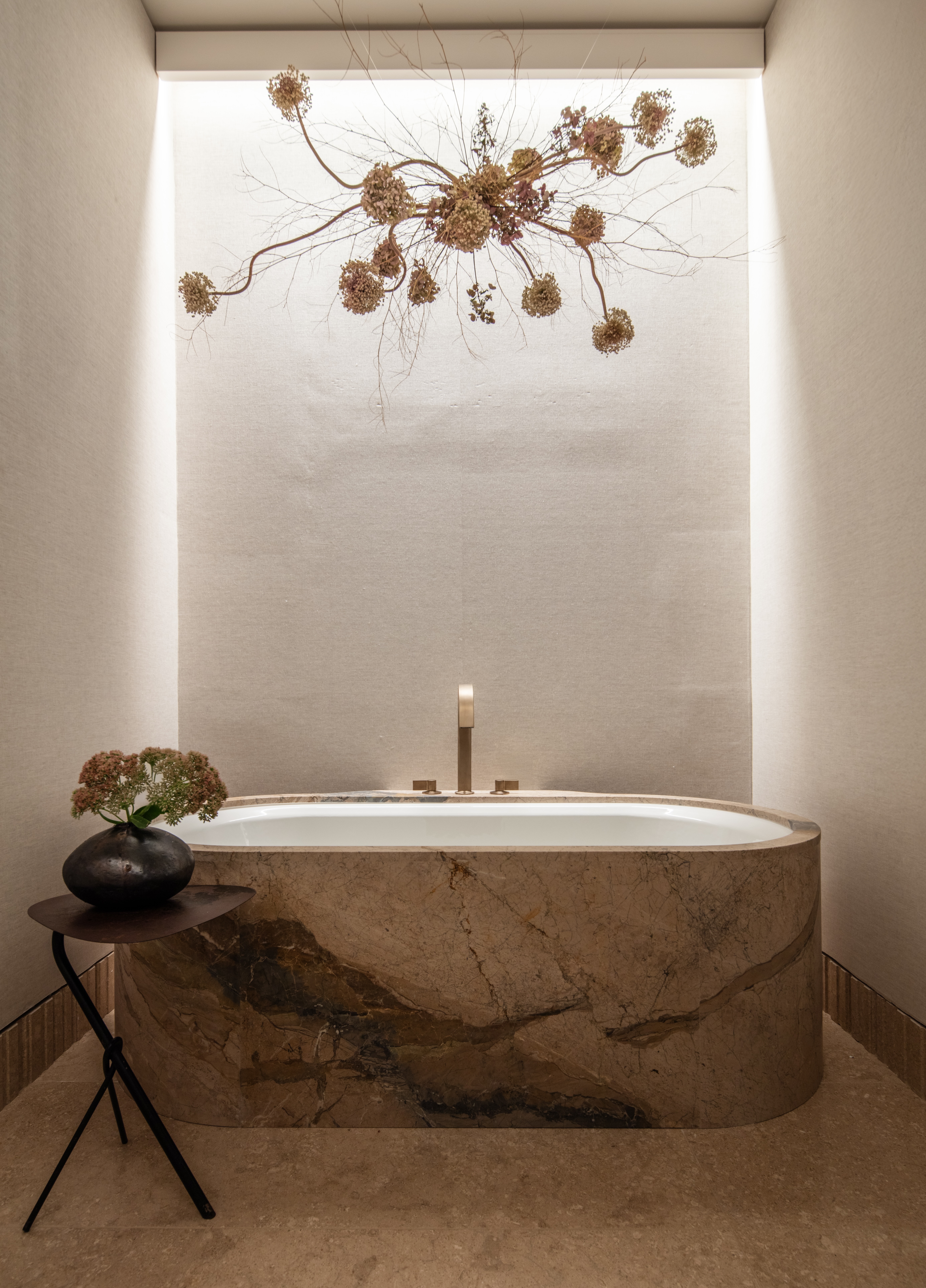 Freestanding marble bathtub with a small side table and vase, under a wall-mounted dried floral arrangement in a softly lit, minimalist bathroom.
