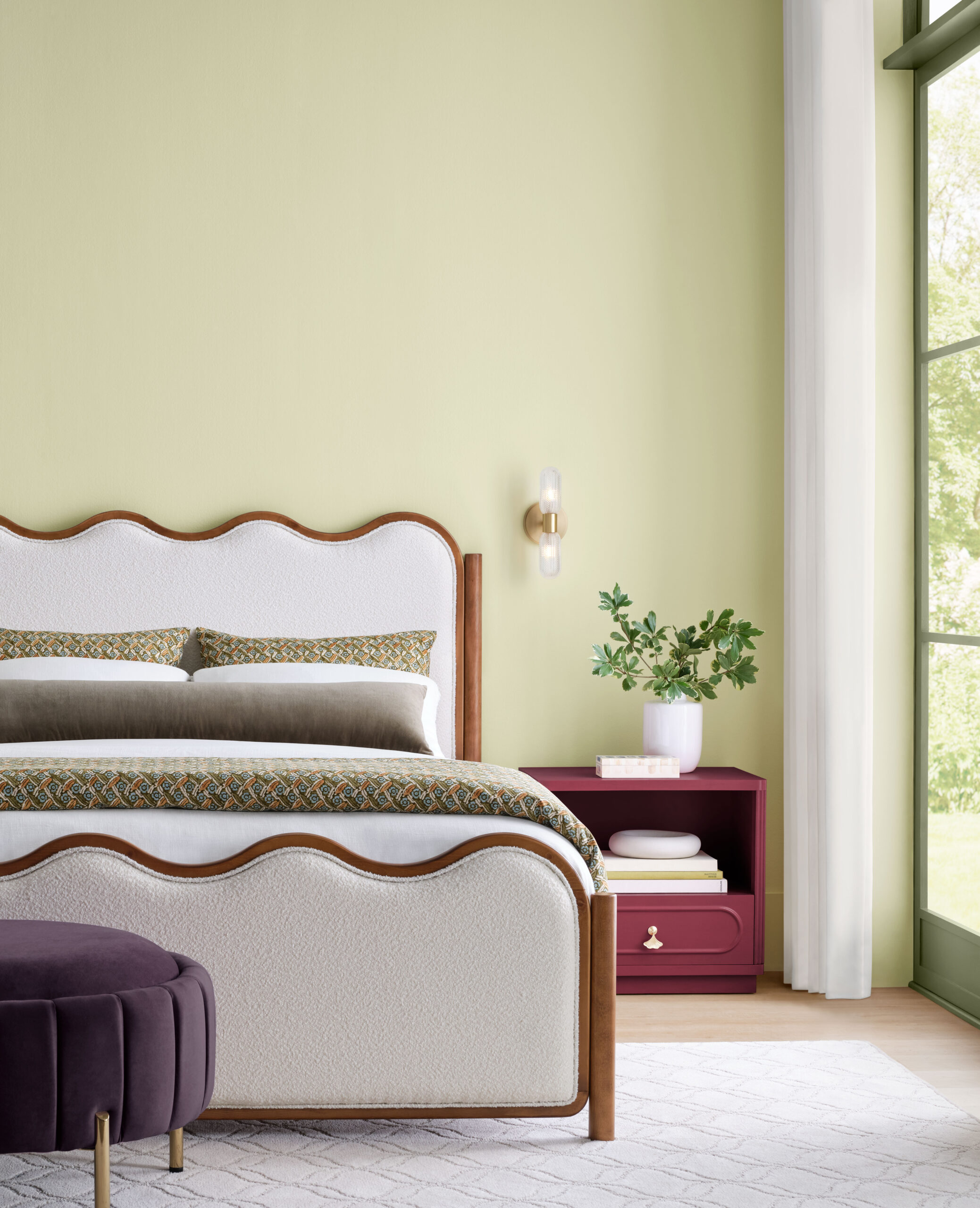 Bedroom with a white and wood bed, patterned bedding, a burgundy nightstand with a potted plant, and a purple ottoman on a geometric rug by a large window.