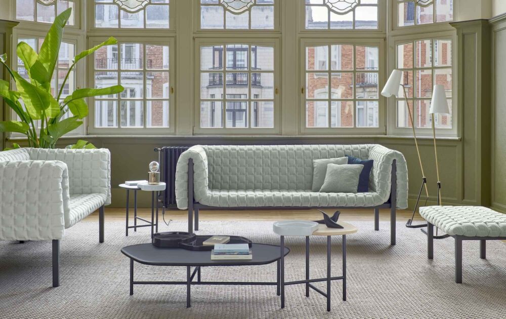 A light green sofa, armchair, and bench with quilted upholstery arranged in a room with large windows, nested coffee tables, and a floor lamp.