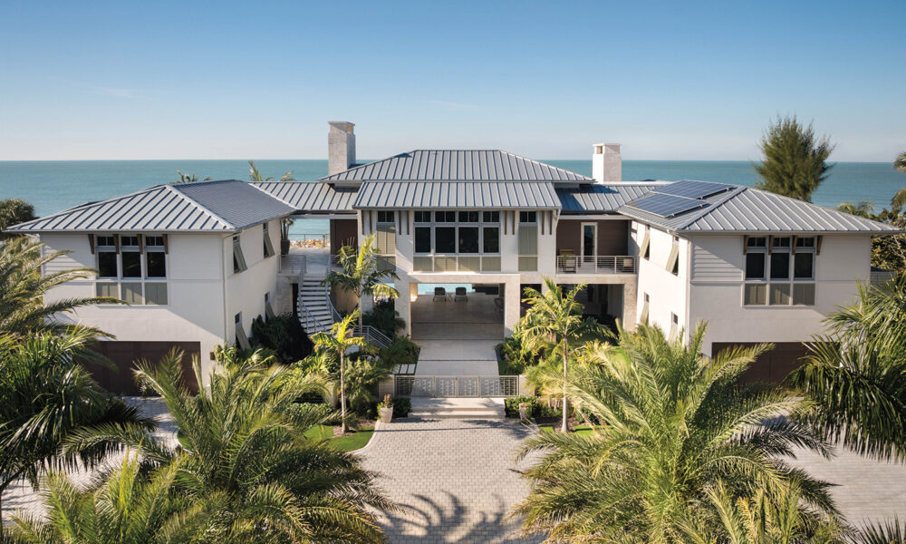 A modern two-story beachfront house with a metal roof and a paved driveway surrounded by palm trees.