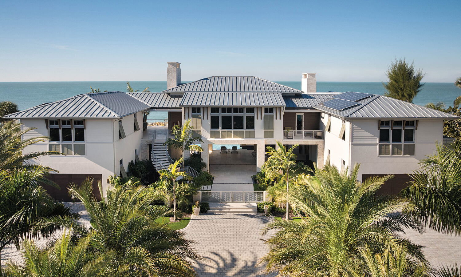 A modern two-story beachfront house with a metal roof and a paved driveway surrounded by palm trees.